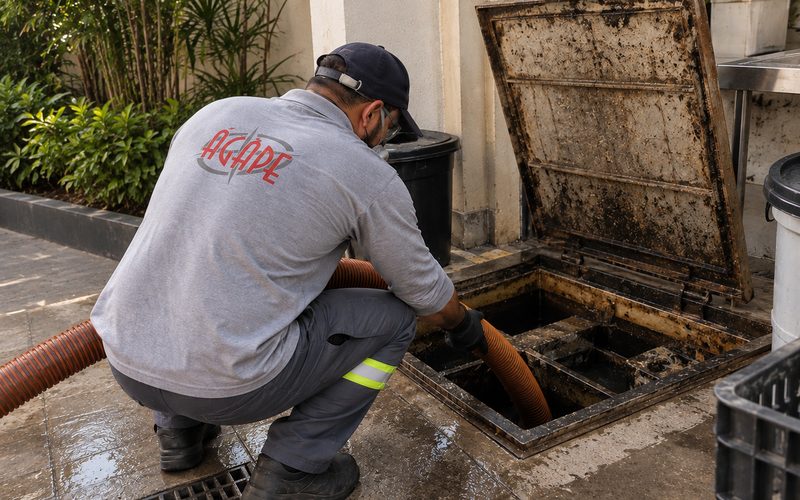 Funcionário uniformizado agachado, com a mangueira de sucção inserida em uma caixa de gordura aberta