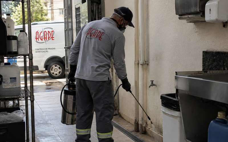 Funcionário uniformizado aplicando produto com pulverizador em um corredor de cozinha, com uma van ao fundo